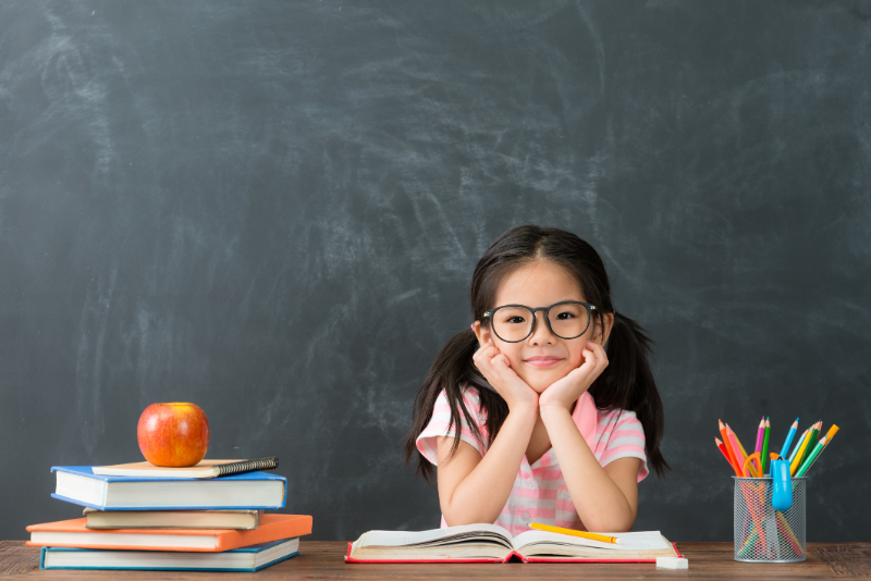Image of a girl in glasses seated at a desk with books and an apple.