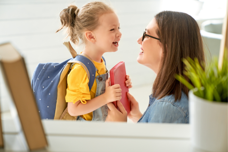 A mother and daughter, wearing a backpack and holding a book, look at each other smiling.