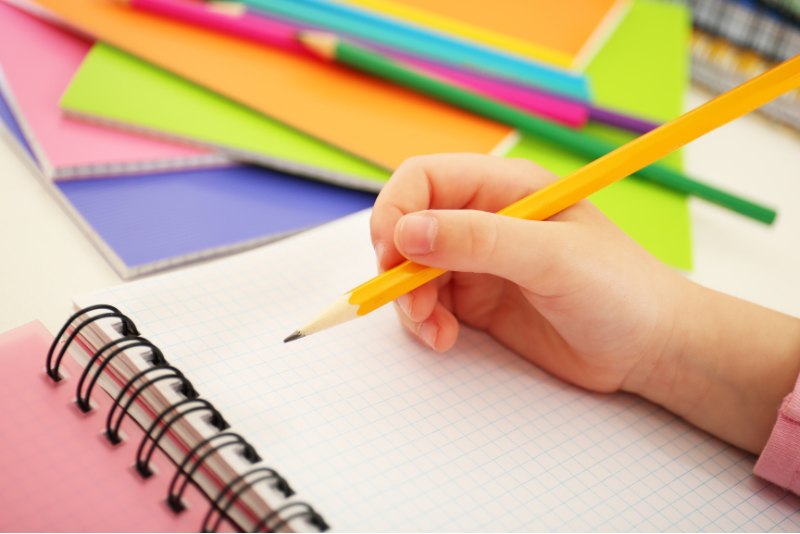 A child's hand holds a pencil above graphing paper with colorful pencils in the background.