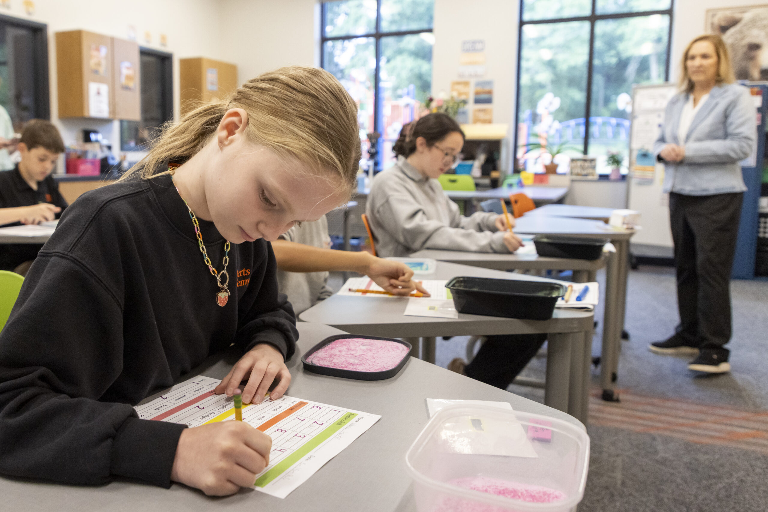 A student at Roberts Academy focuses on writing during a structured literacy lesson using the Orton-Gillingham approach. She sits at her desk with a colorful phonics chart and a tactile tray filled with pink sand, engaging in multisensory learning designed to support dyslexic students. Other students and a teacher are visible in the background of the bright, welcoming classroom.