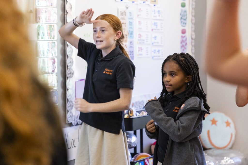 Two elementary students at Roberts Academy participate in an Orton-Gillingham classroom activity that incorporates movement and gestures to reinforce learning. The girls, dressed in school uniforms, stand near a whiteboard covered in phonics patterns and charts, actively engaging in a multisensory literacy lesson designed to support students with dyslexia.
