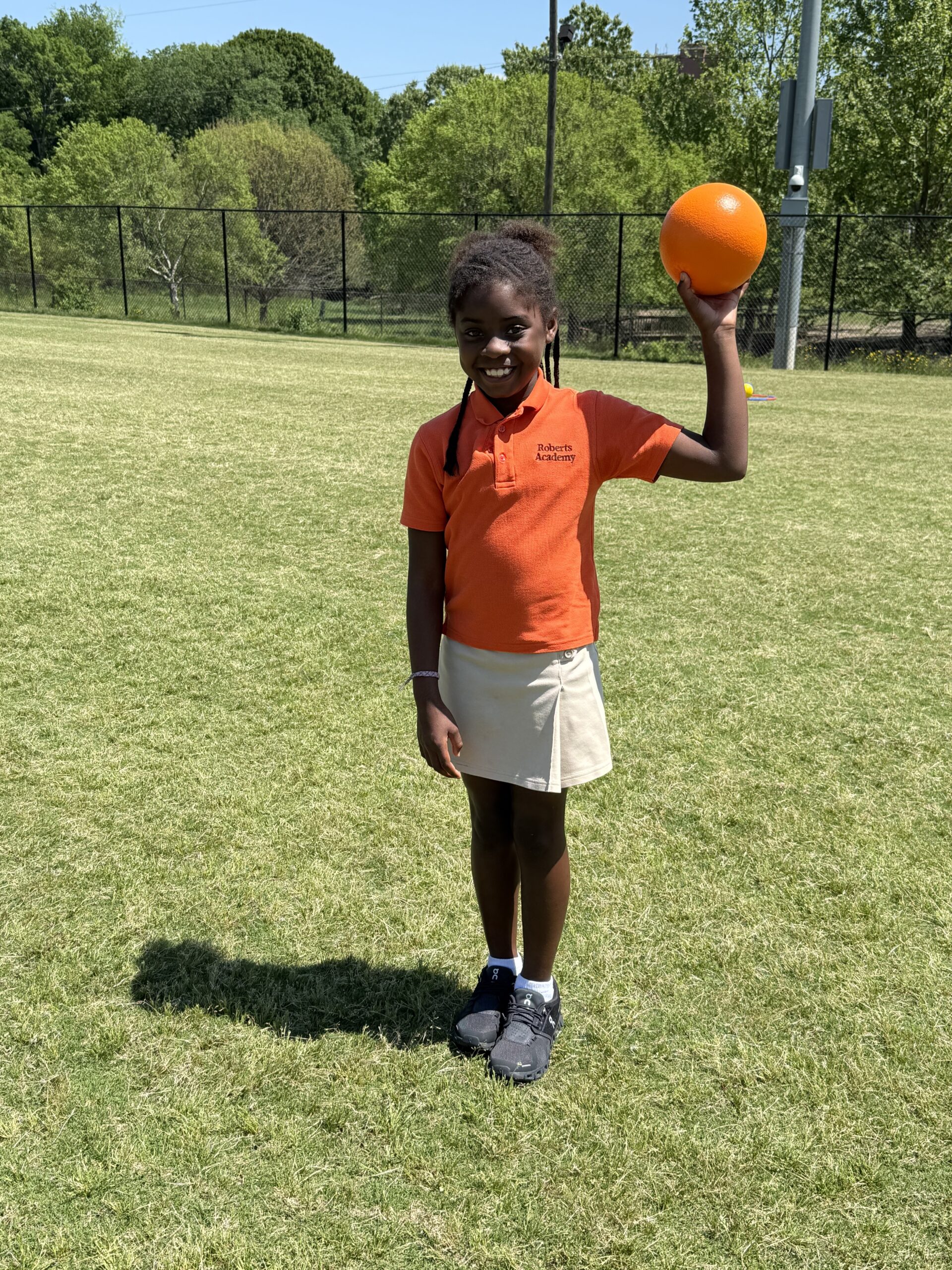 Smiling Roberts Academy student in an orange uniform polo and khaki skirt stands on a sunny field holding an orange ball, enjoying PE on campus surrounded by green trees and a black chain-link fence