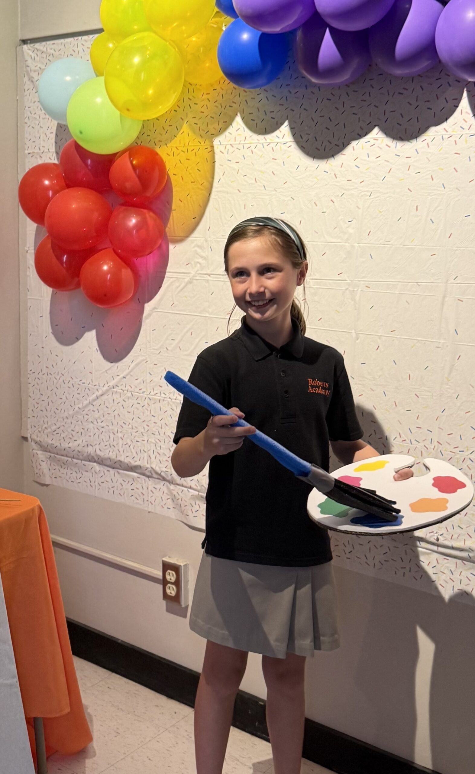 Smiling Roberts Academy student in school uniform holds a colorful artist palette and oversized paintbrush, standing in front of a rainbow balloon arch and sprinkle-patterned backdrop during a creative arts activity.