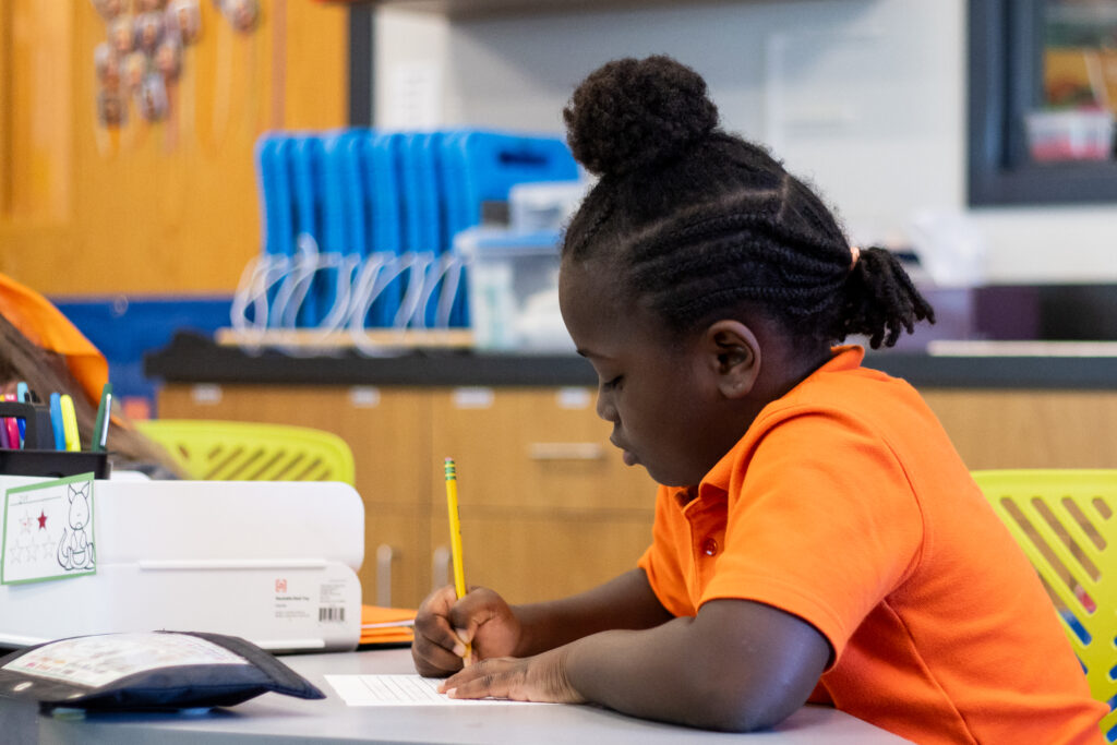 A Roberts Academy 2nd grader in an orange polo shirt sits at a desk and writes on a sheet of paper