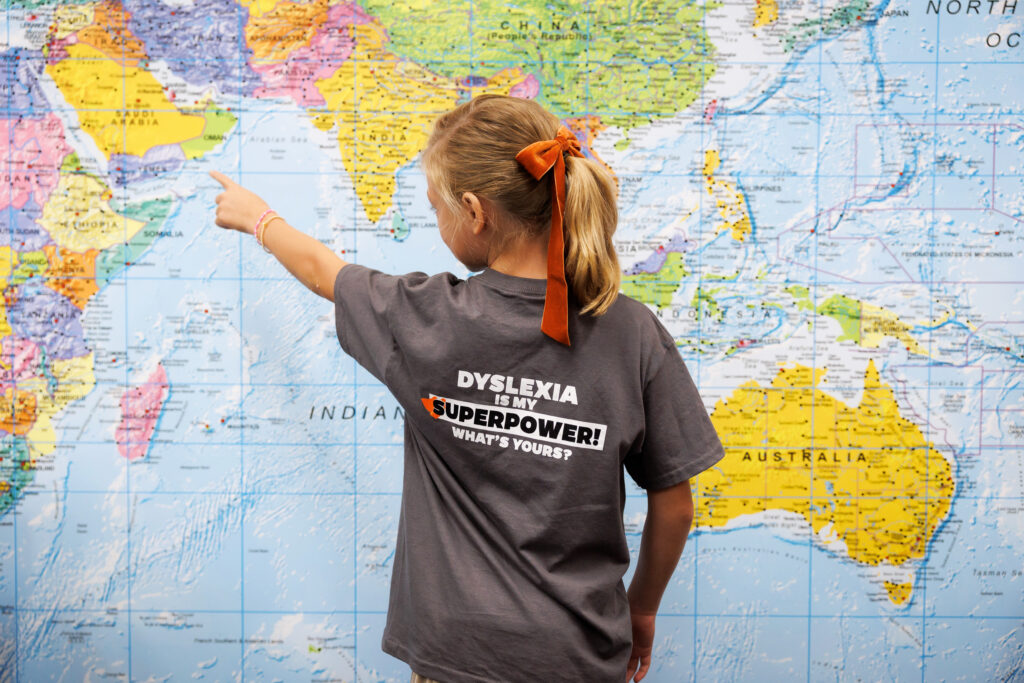 A Roberts Academy at Mercer University Student points to a a country on a large wall-map. She is wearing an orange bow and a gray t-shirt that reads, "Dyslexia is my superpower. What's yours?