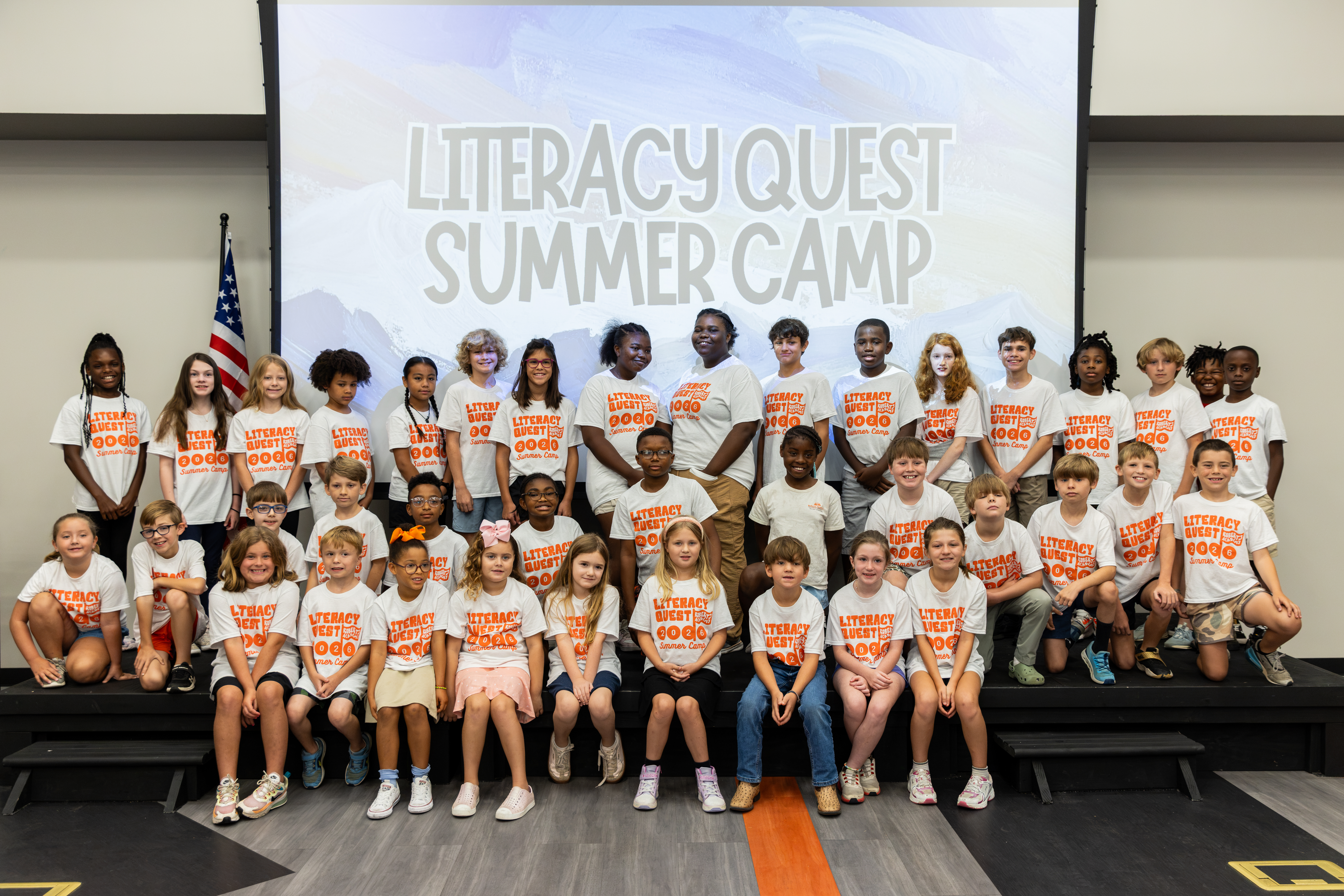 Group of about 40 elementary-age children wearing white "Literacy Quest 2026 Summer Camp" T-shirts, posed together on stage in front of a Literacy Quest Summer Camp projection screen at Roberts Academy at Mercer University.