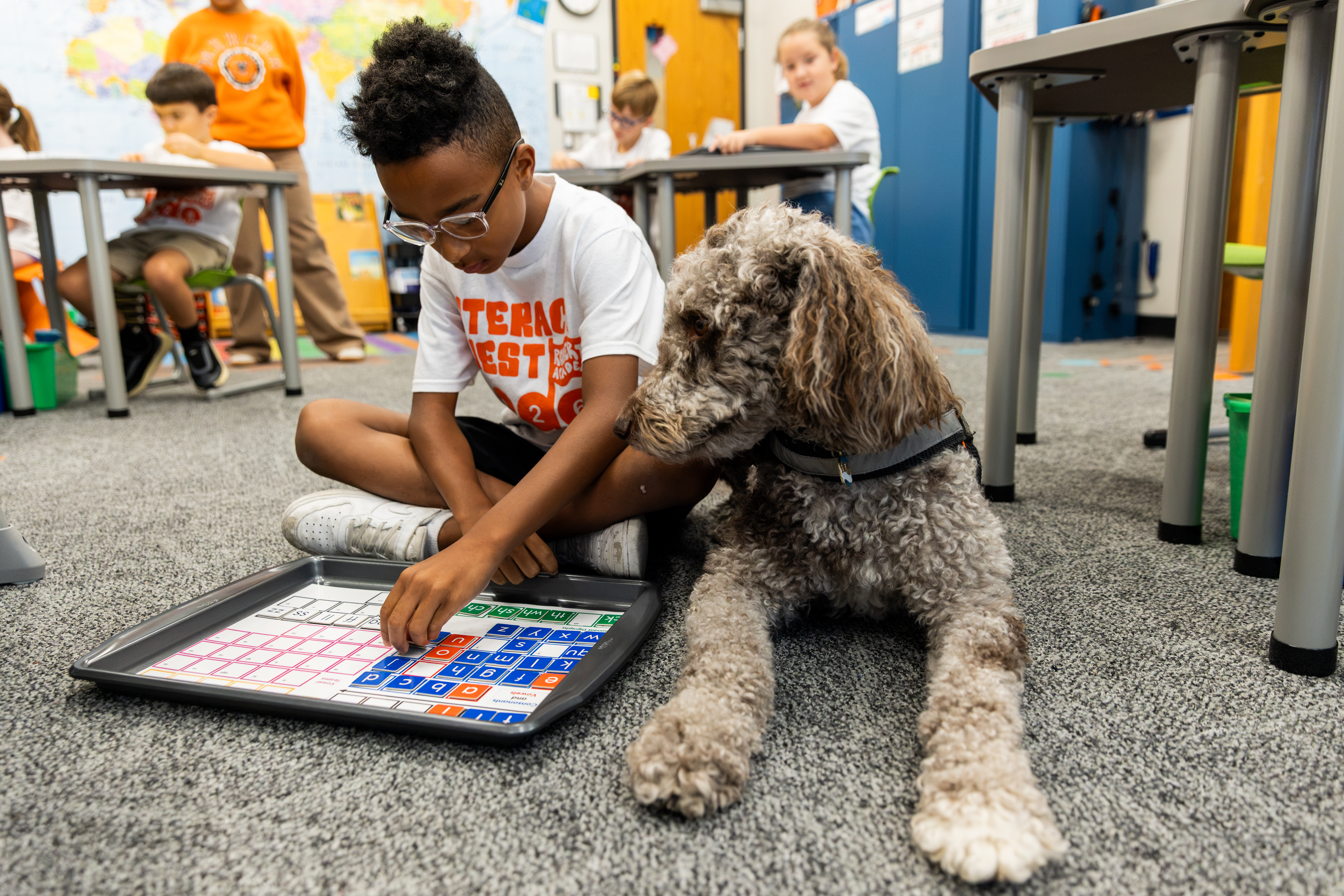 A young student Literacy Quest Summer Camp student sits cross-legged on a classroom floor at Roberts Academy summer camp, arranging letter tiles on a magnetic phonics tray while a curly-haired therapy dog rests beside him, with classmates working at desks in the background.
