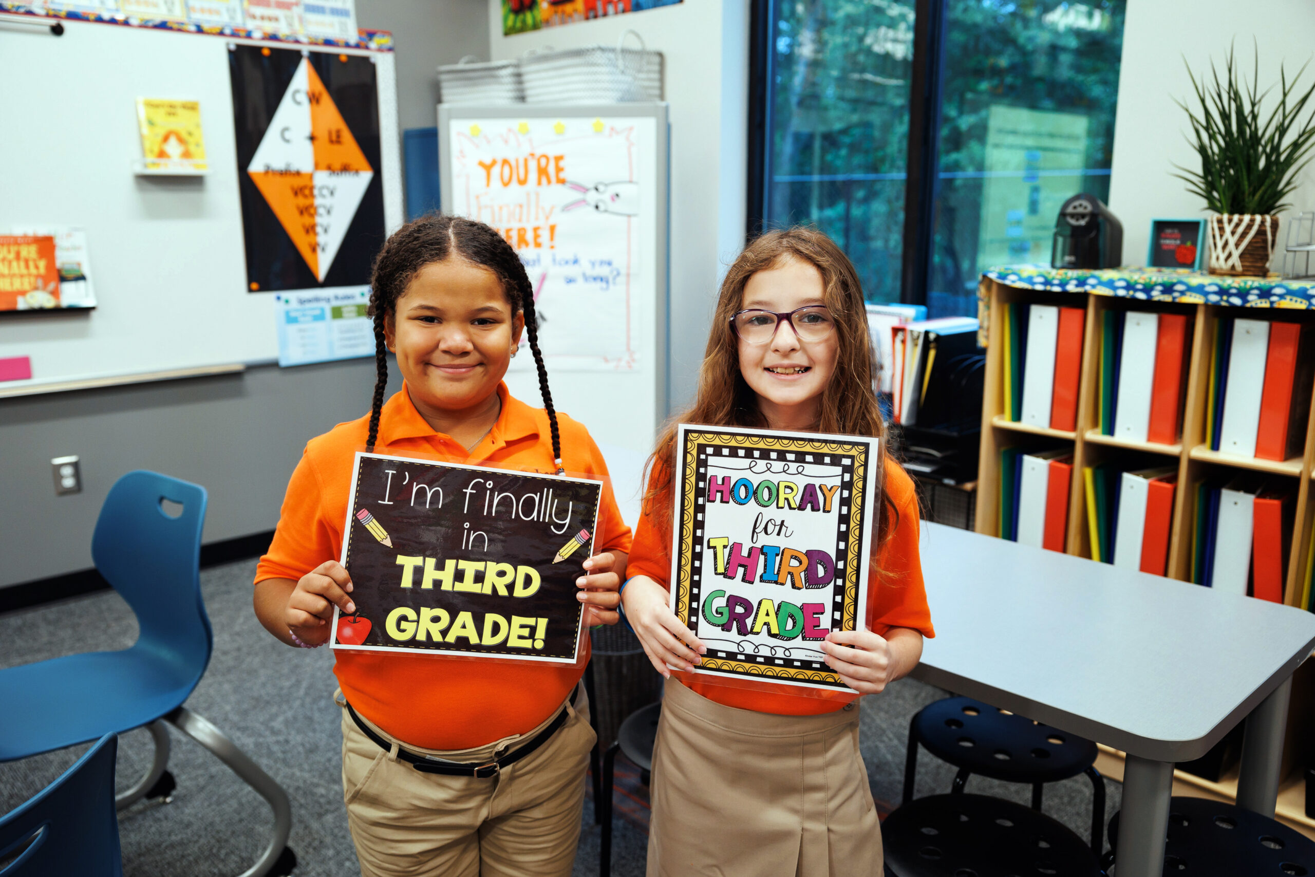 Two smiling third-grade students in orange Roberts Academy polos hold "I'm finally in Third Grade!" and "Hooray for Third Grade" signs on the first day of school in a bright, decorated classroom.